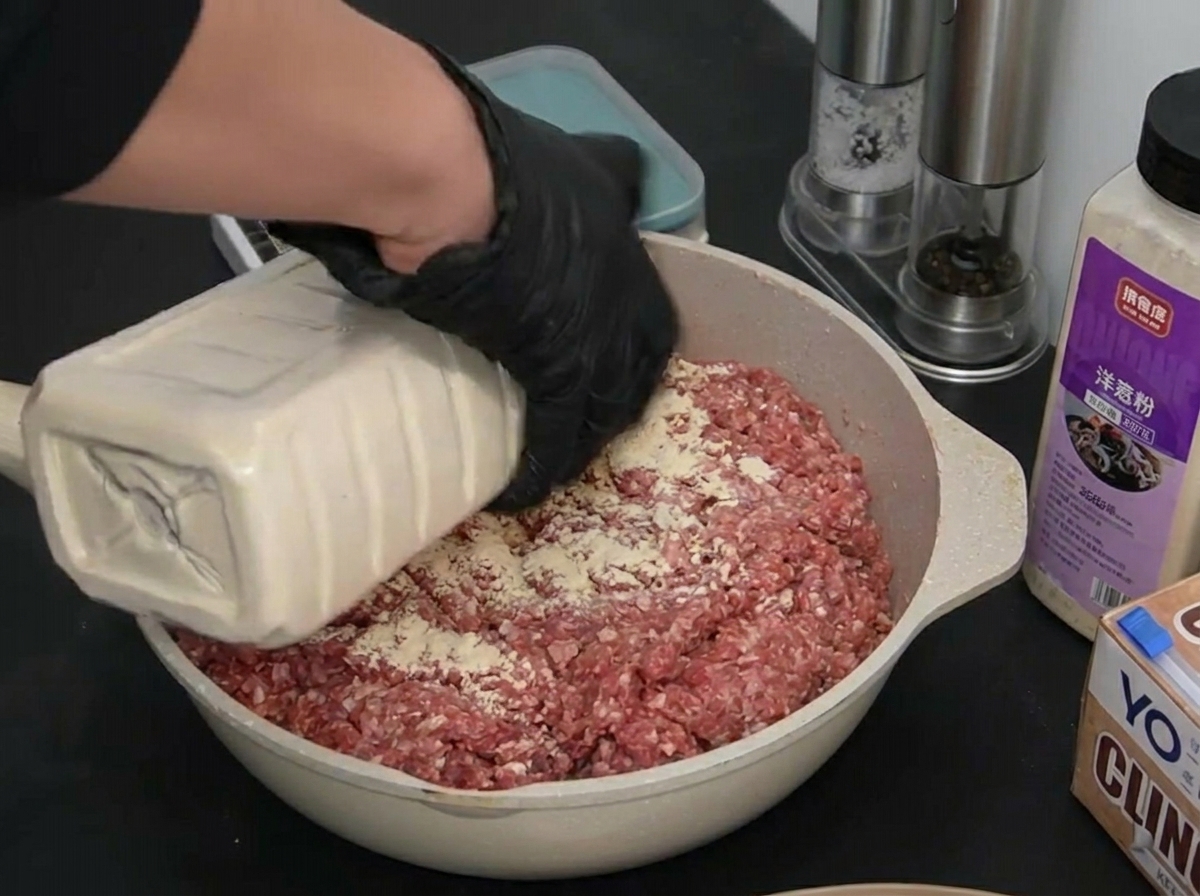 Una persona con guantes negros vertiendo ajo en polvo blanco desde un recipiente grande sobre la carne picada.