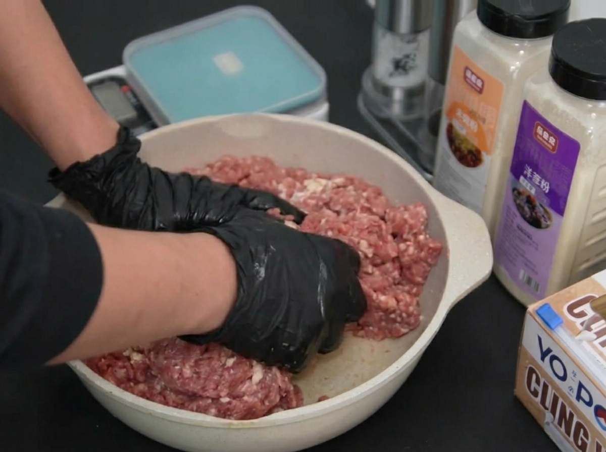 Dos manos con guantes presionando y mezclando la carne picada dentro de una bandeja.