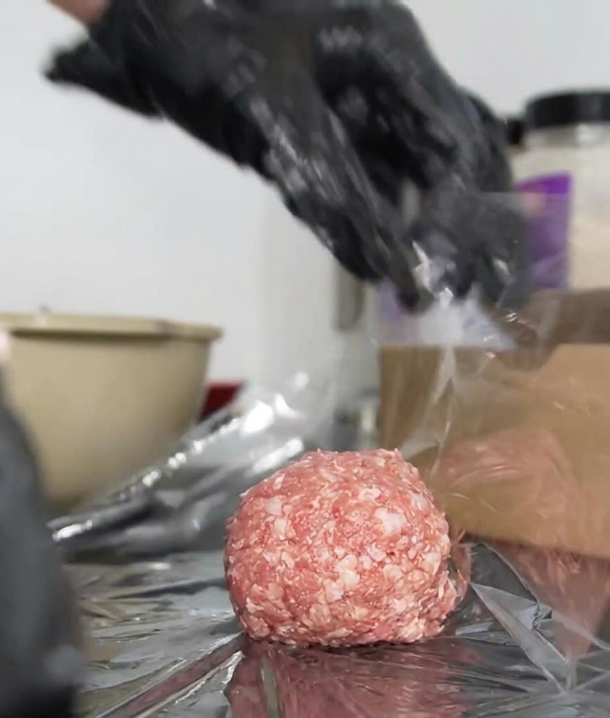 Manos con guantes girando con fuerza el film transparente alrededor de una hamburguesa para crear un paquete sellado.