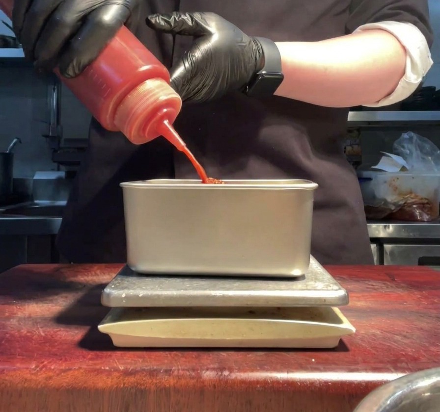 Manos con guantes vertiendo salsa sriracha roja desde una botella a un contenedor de metal.