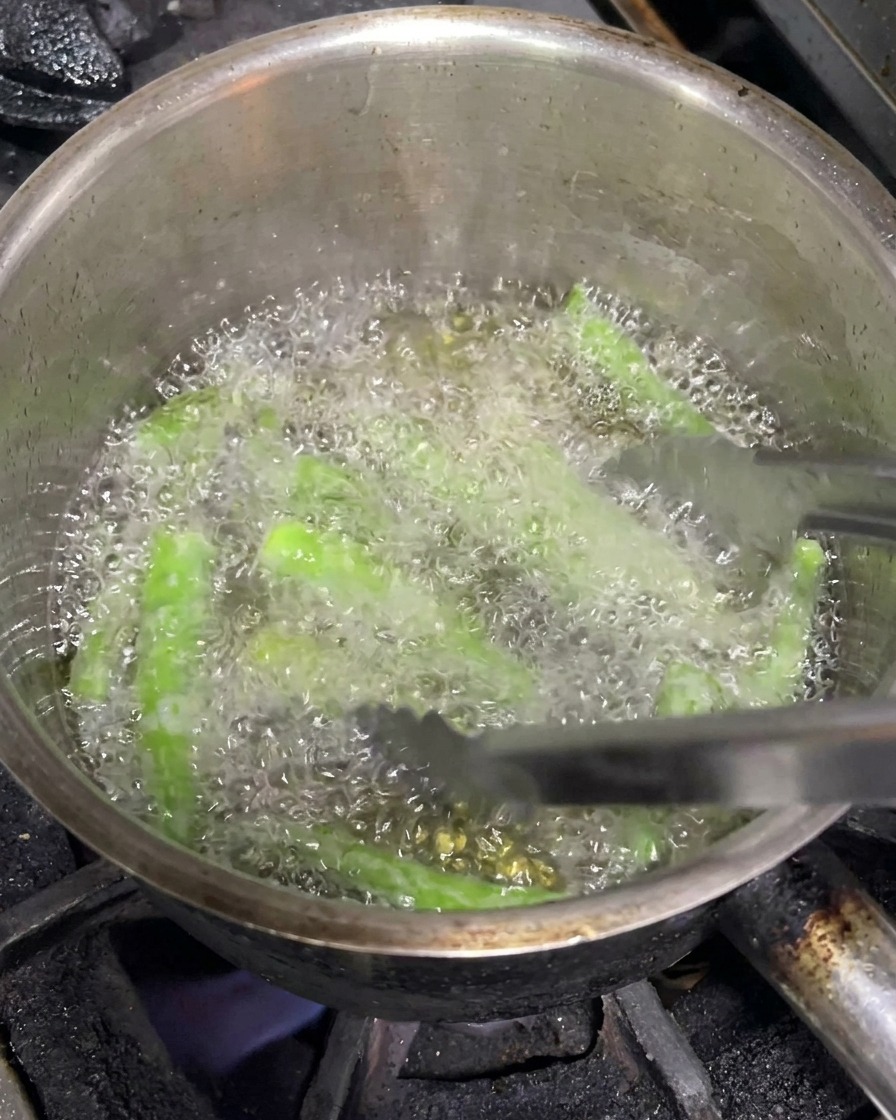 Pinzas metálicas moviendo chiles verdes enteros mientras se fríen en aceite caliente.