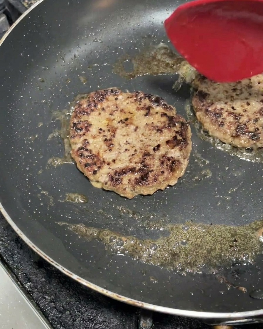 Dos piezas de carne de hamburguesa cocinándose en una plancha caliente con una espátula roja encima.