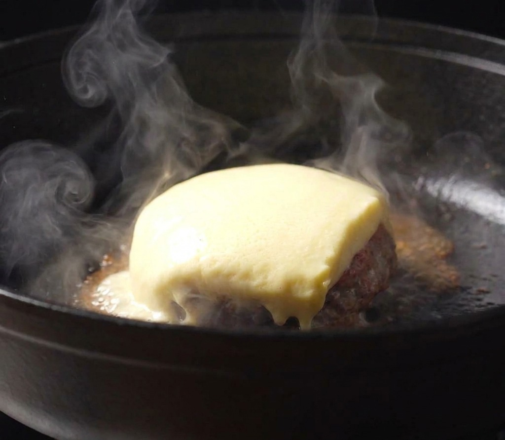 Loncha de queso amarillo fundiéndose sobre una hamburguesa con salsa en una sartén de hierro.