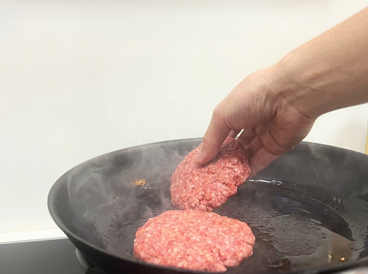 Una mano colocando dos porciones de carne de ternera cruda en una sartén negra muy caliente.