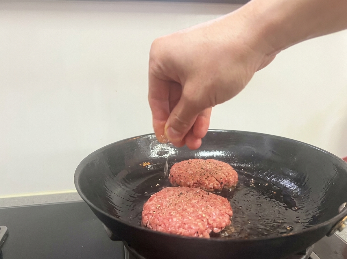 Una mano espolvoreando sal y pimienta negra sobre dos piezas de carne sellándose en una sartén.