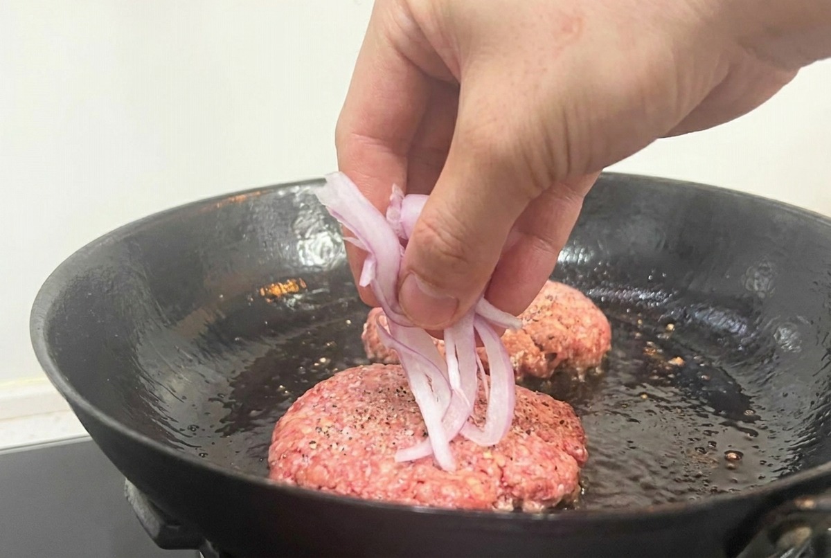 Una mano dejando caer rodajas de cebolla roja sobre la carne cruda que se cocina en una sartén negra.