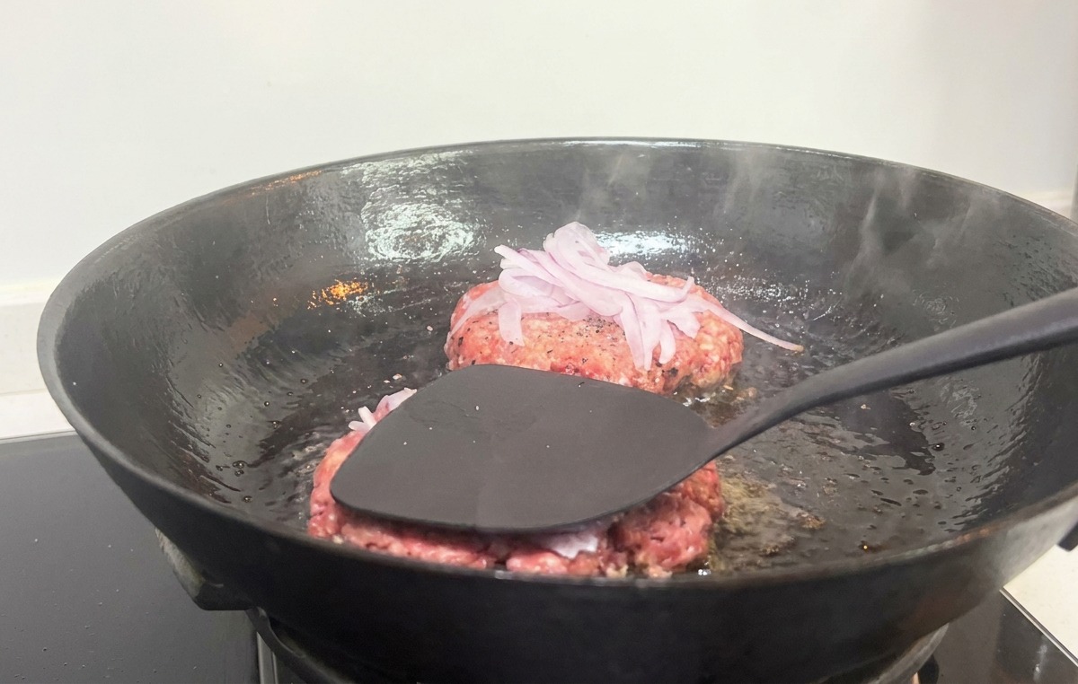 Una espátula negra presionando firmemente una hamburguesa con cebolla roja en una sartén de hierro fundido caliente.