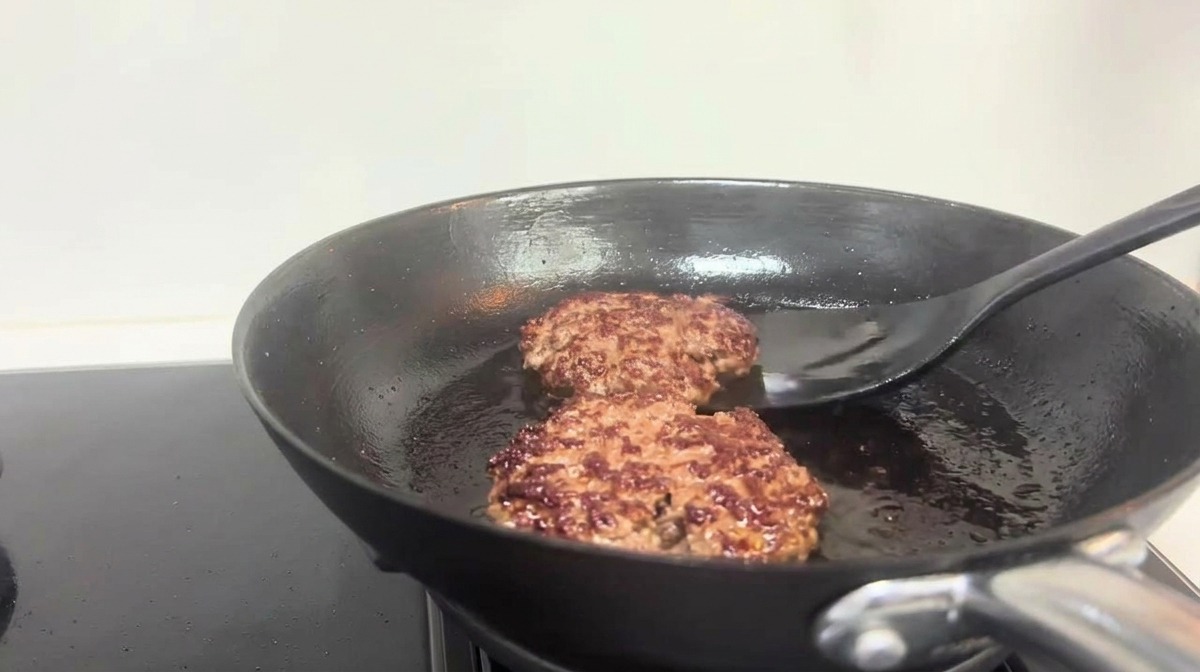 Una espátula negra dando la vuelta a hamburguesas de ternera doradas en una sartén.