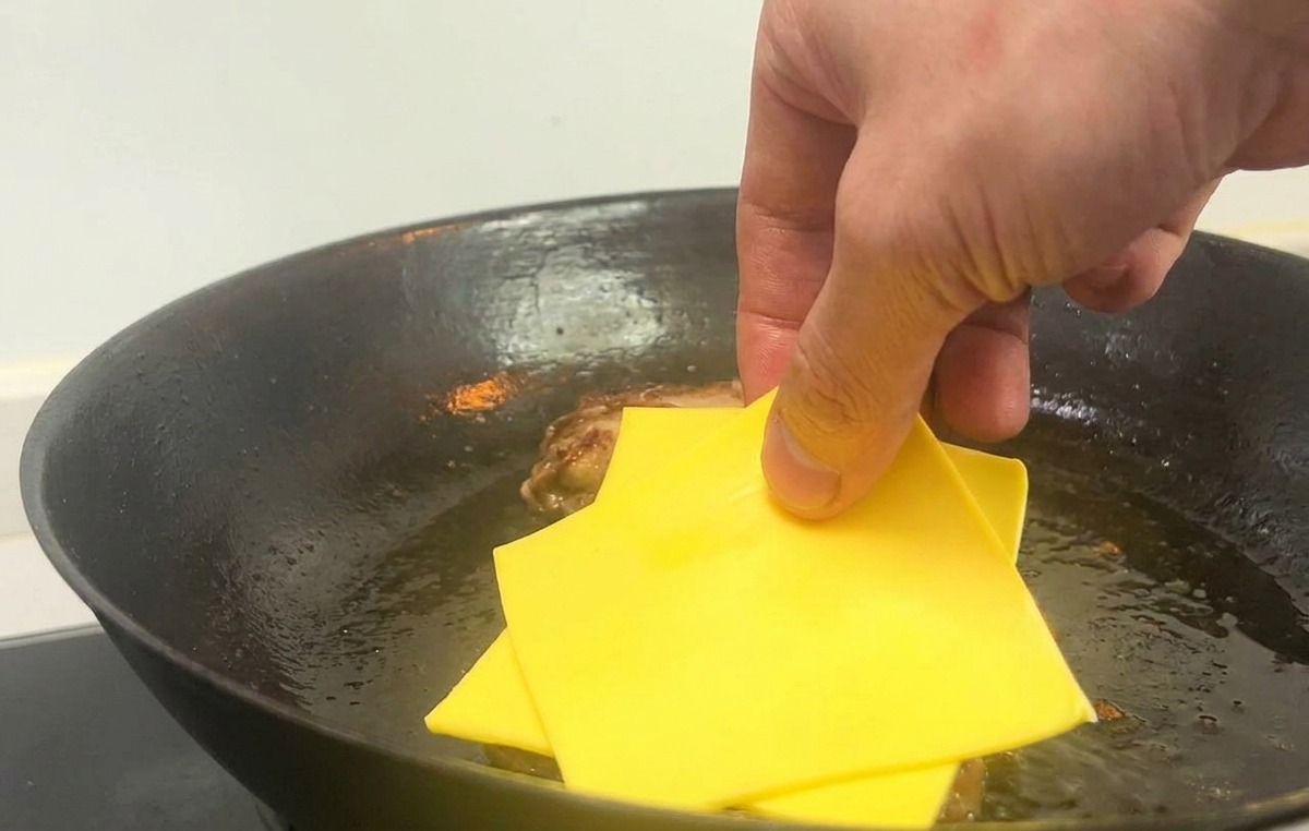 Una mano colocando dos lonchas cuadradas de queso amarillo sobre hamburguesas cocinadas en una sartén caliente.
