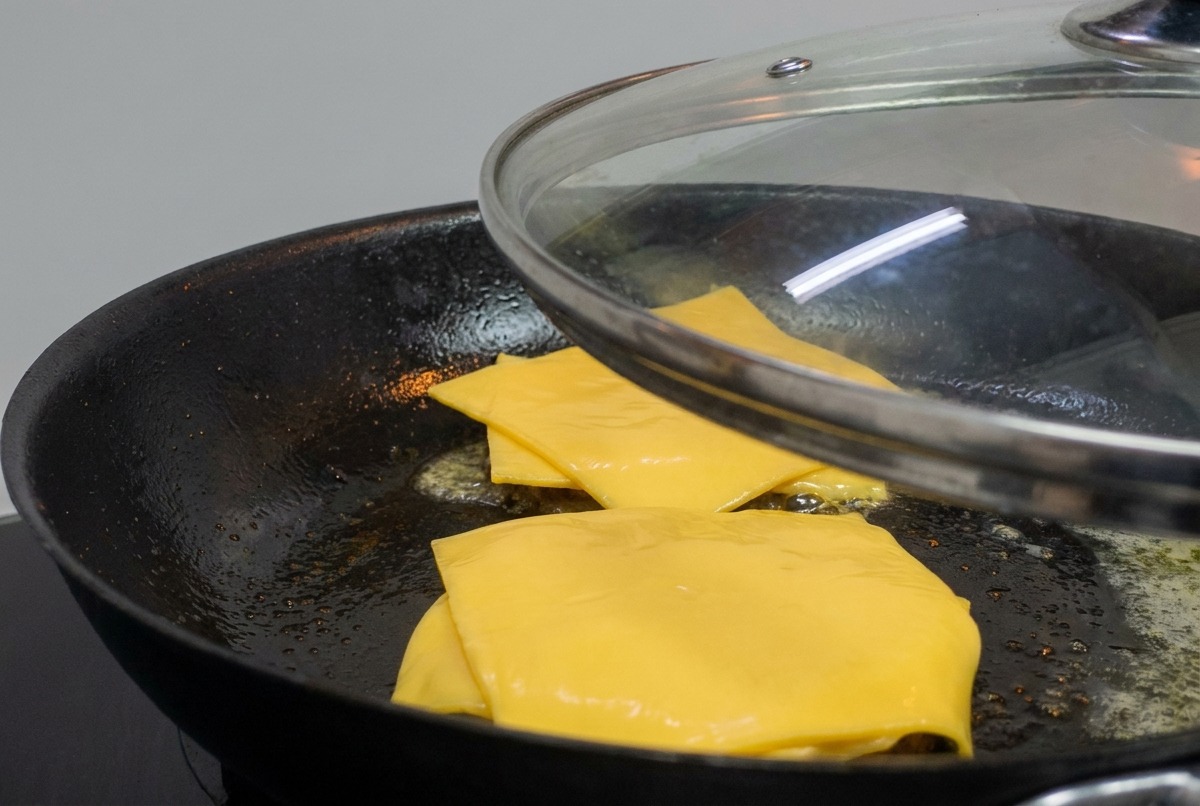 Una tapa de cristal colocándose sobre una sartén con hamburguesas cubiertas de queso amarillo fundido.