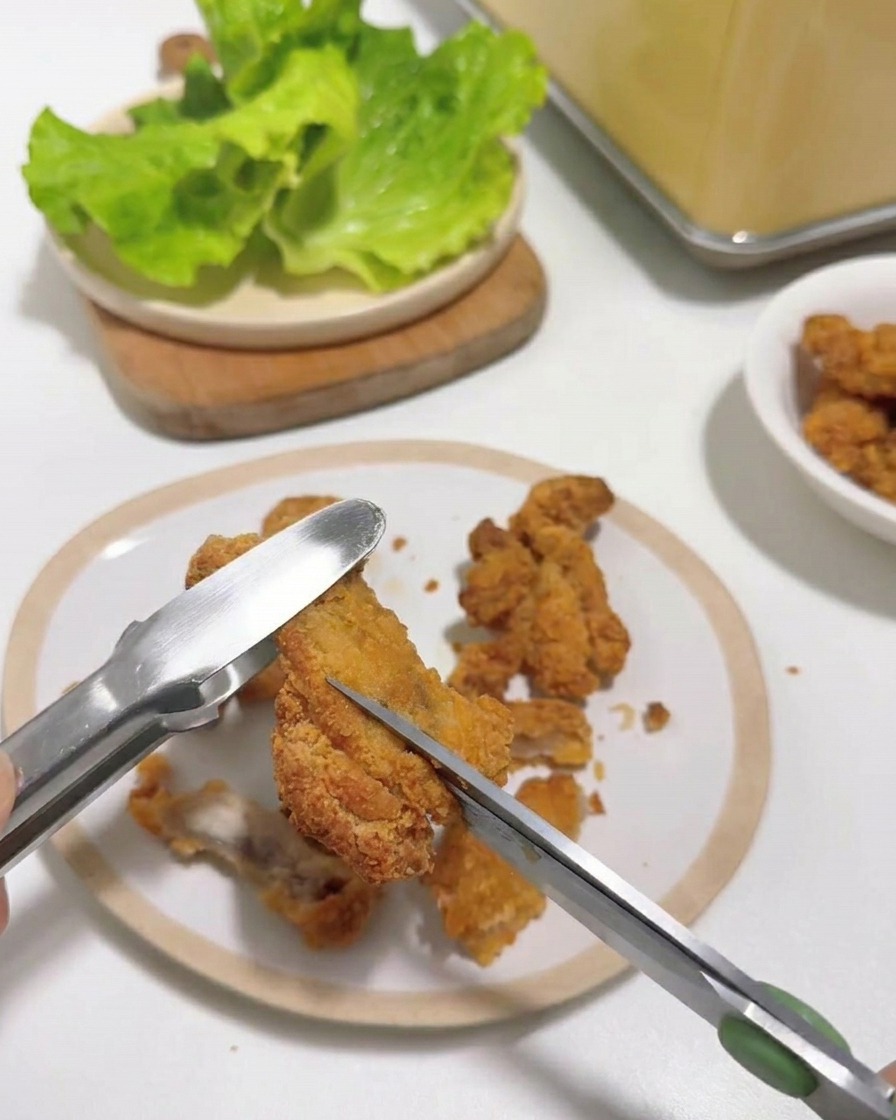 Primer plano de unas tijeras de cocina cortando pollo frito caliente sostenido con pinzas.