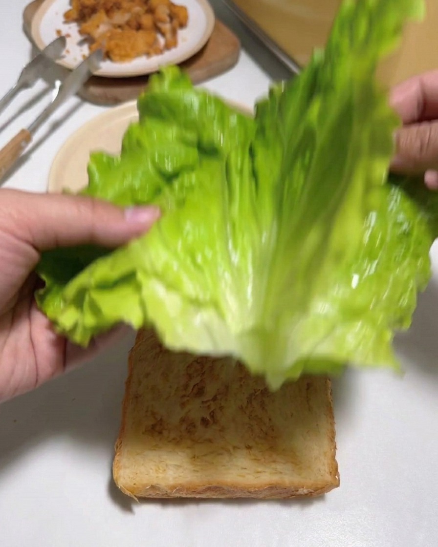 Manos colocando hojas verdes de lechuga fresca sobre una rebanada de pan tostado.