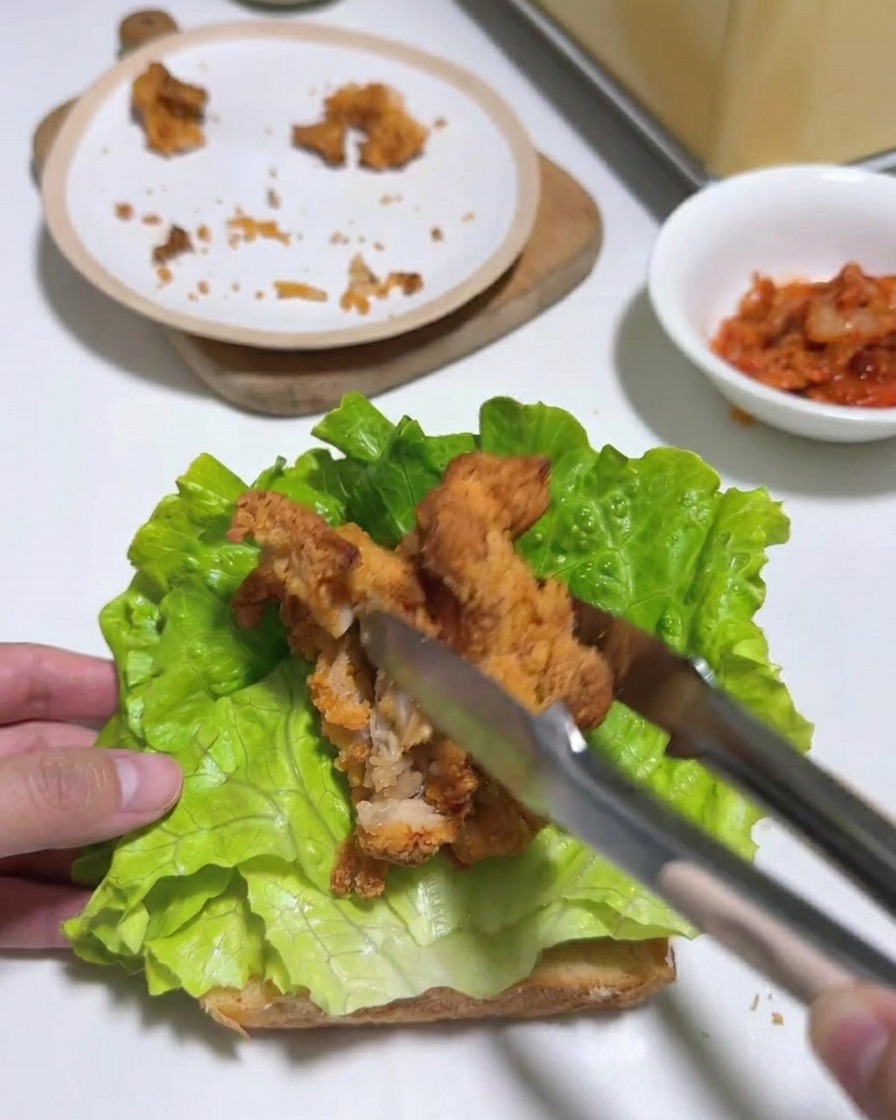 Pinzas colocando tiras de pollo frito sobre la cama de lechuga en el pan tostado.
