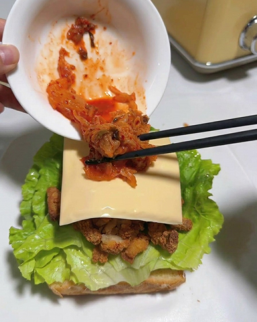 Palillos pasando kimchi rojo picado de un cuenco al sándwich sobre la loncha de queso.