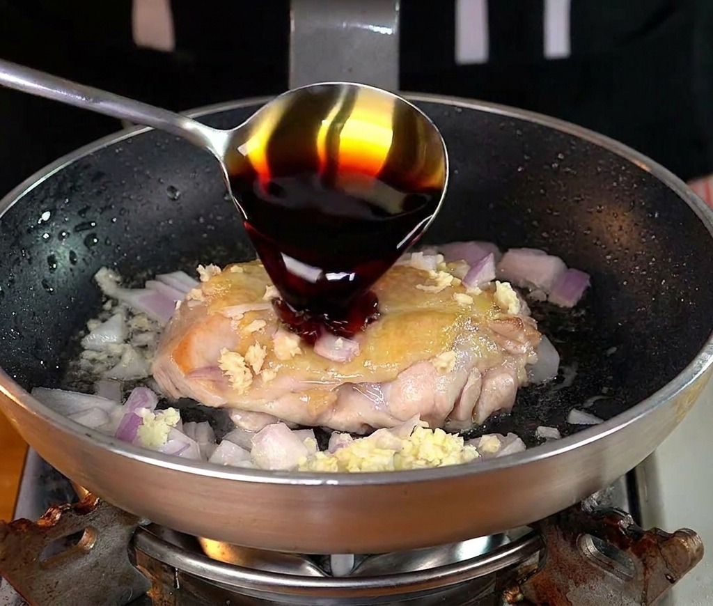 Una cuchara vertiendo salsa de soja sobre un muslo de pollo en la sartén.