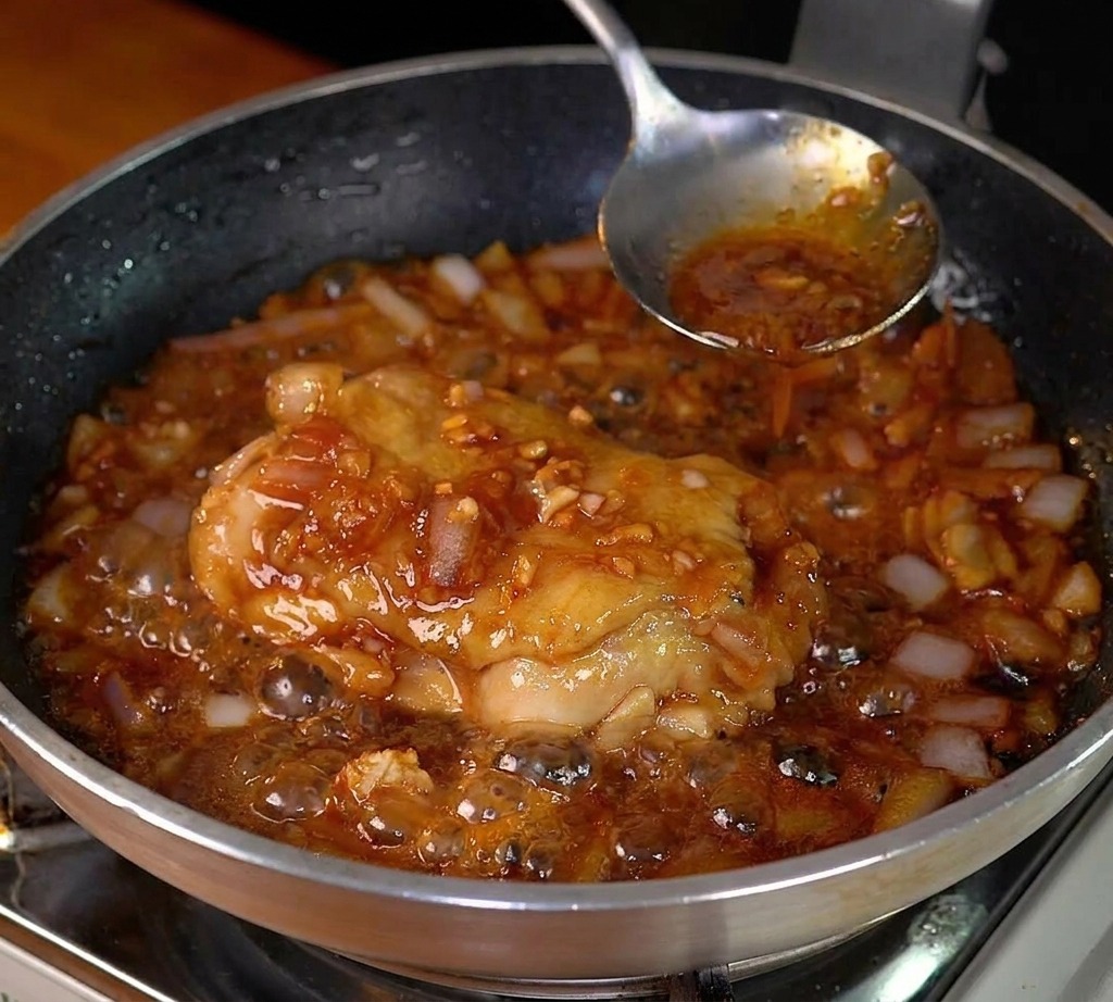 Pollo cocinándose en una salsa oscura y burbujeante en una sartén de acero.