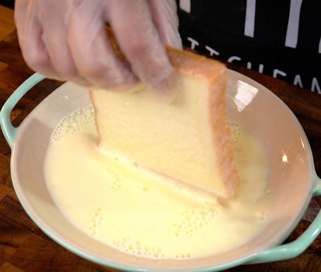 Una mano sumergiendo una rebanada gruesa de pan blanco en la mezcla de huevo.