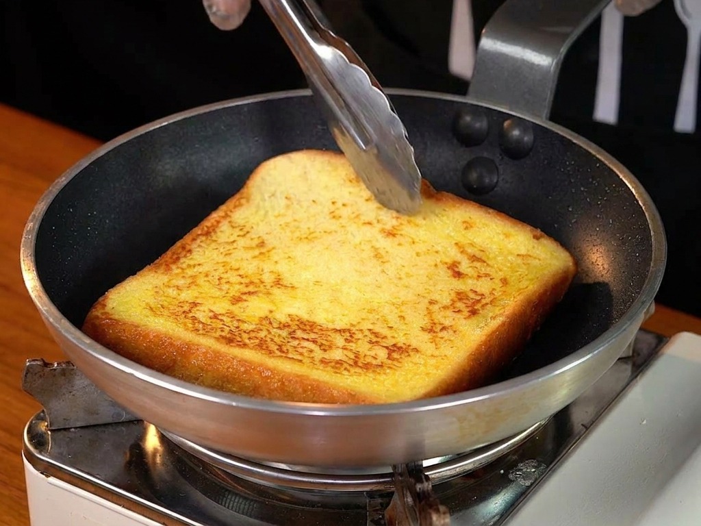 Una rebanada de tostada francesa dorada siendo volteada en una sartén con pinzas.