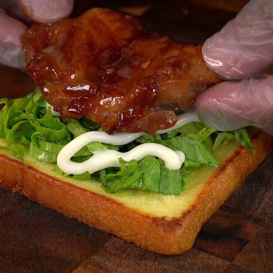 Manos colocando un muslo de pollo brillante sobre una base de pan con lechuga y mayonesa.