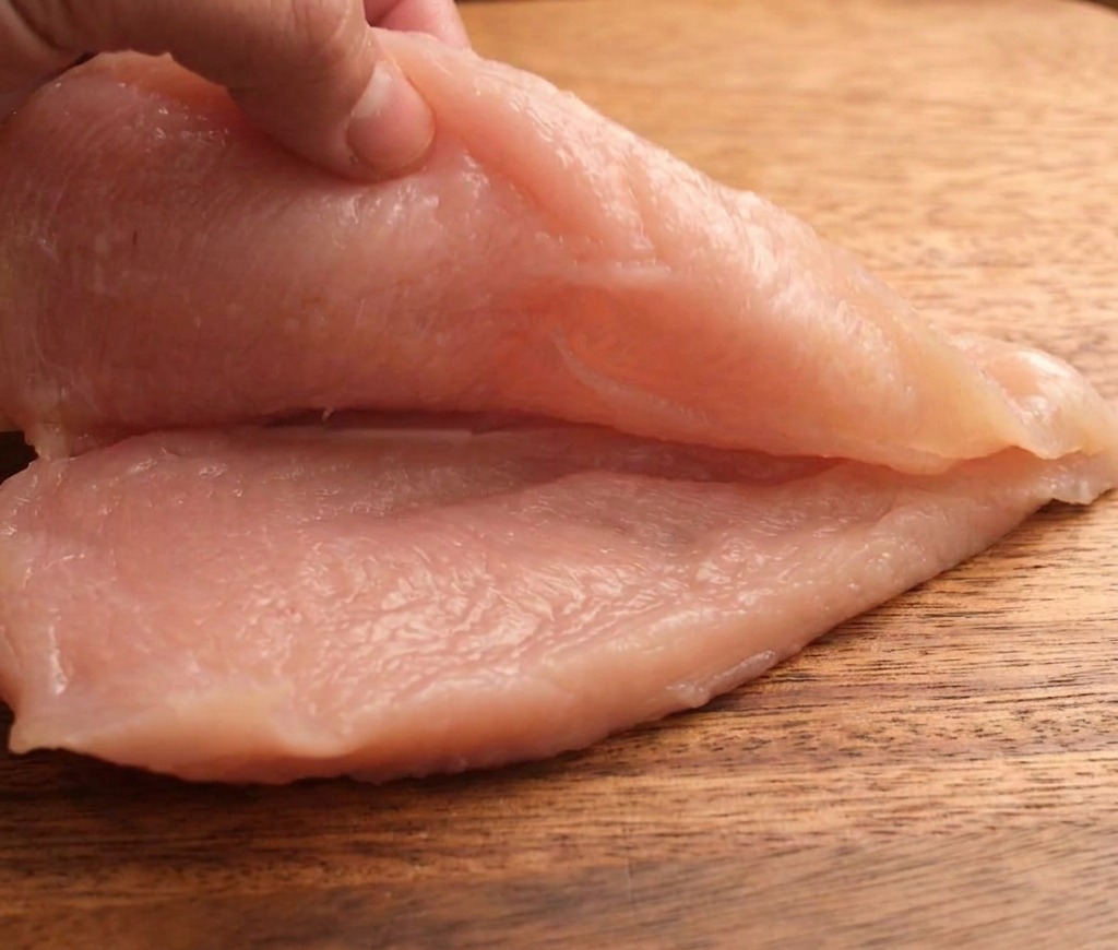 Una mano cortando una pechuga de pollo cruda horizontalmente en dos piezas finas sobre una tabla de madera.
