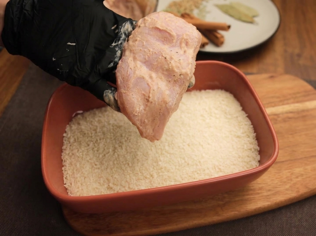 Una mano con guante sosteniendo un filete de pollo marinado sobre una bandeja con pan rallado blanco.