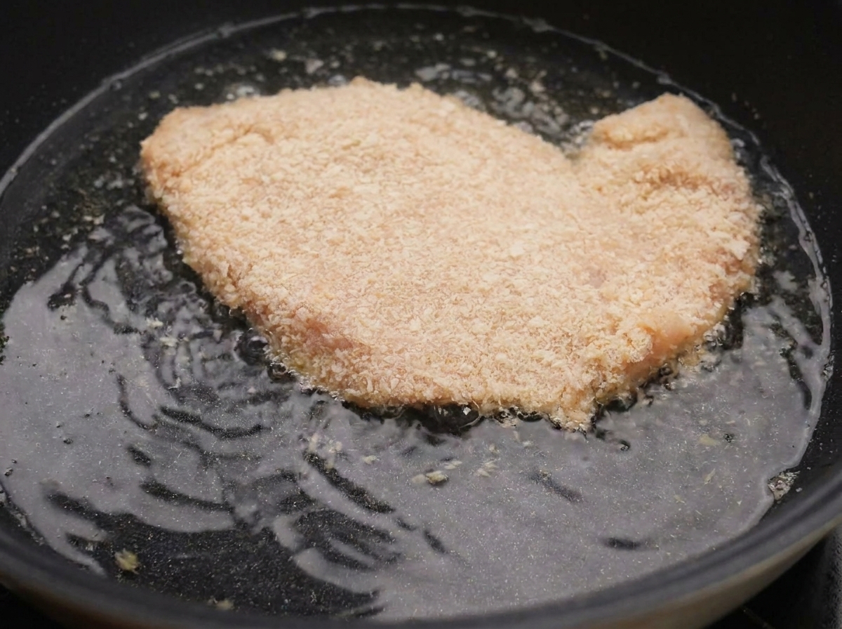 Una pechuga de pollo empanada burbujeando en aceite caliente dentro de una sartén negra.