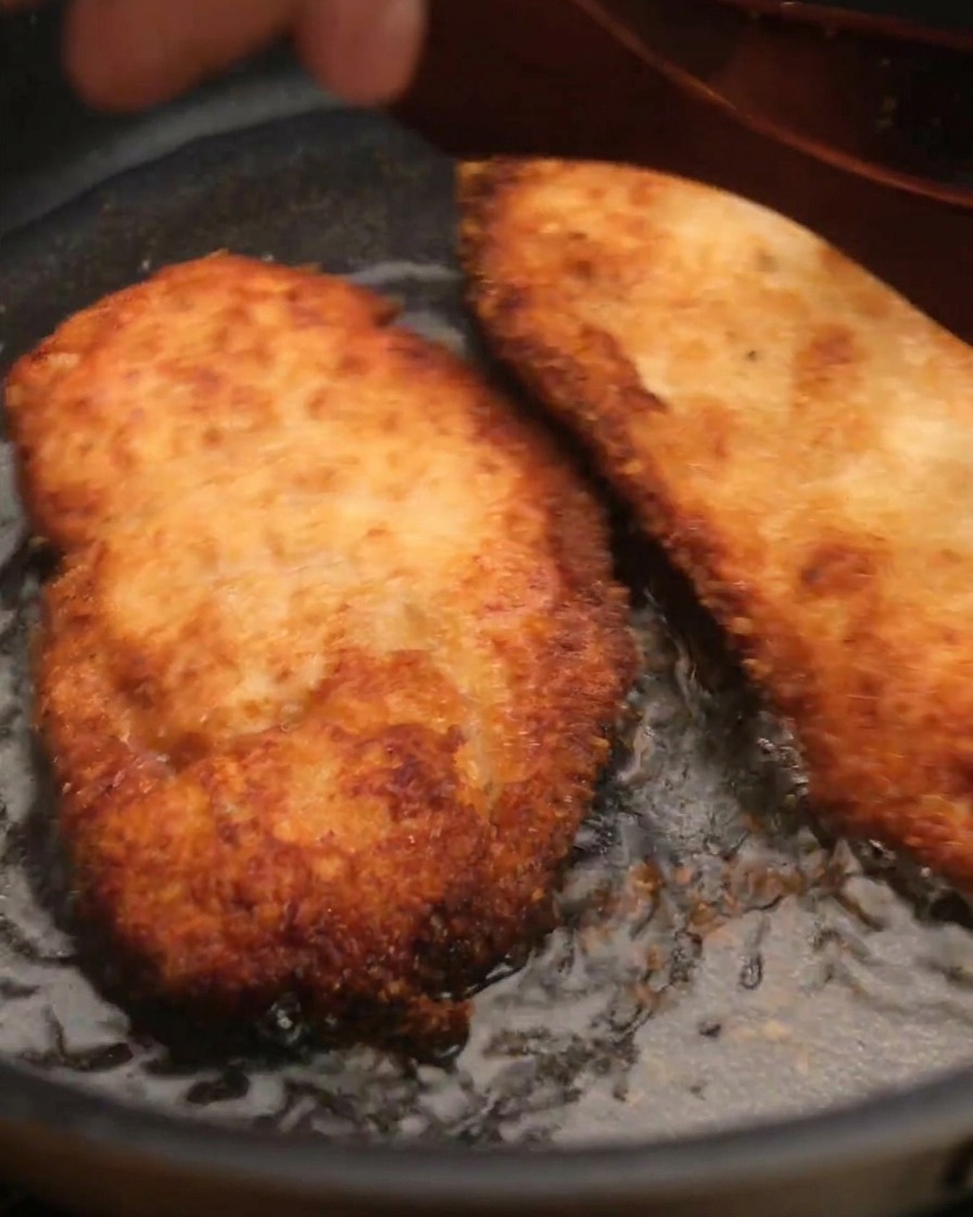 Un trozo de pollo frito crujiente siendo levantado de una sartén con aceite usando una espátula de madera.