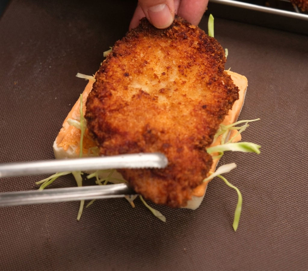 Una mano y pinzas metálicas colocando un filete de pollo frito sobre una rebanada de pan con salsa y col.