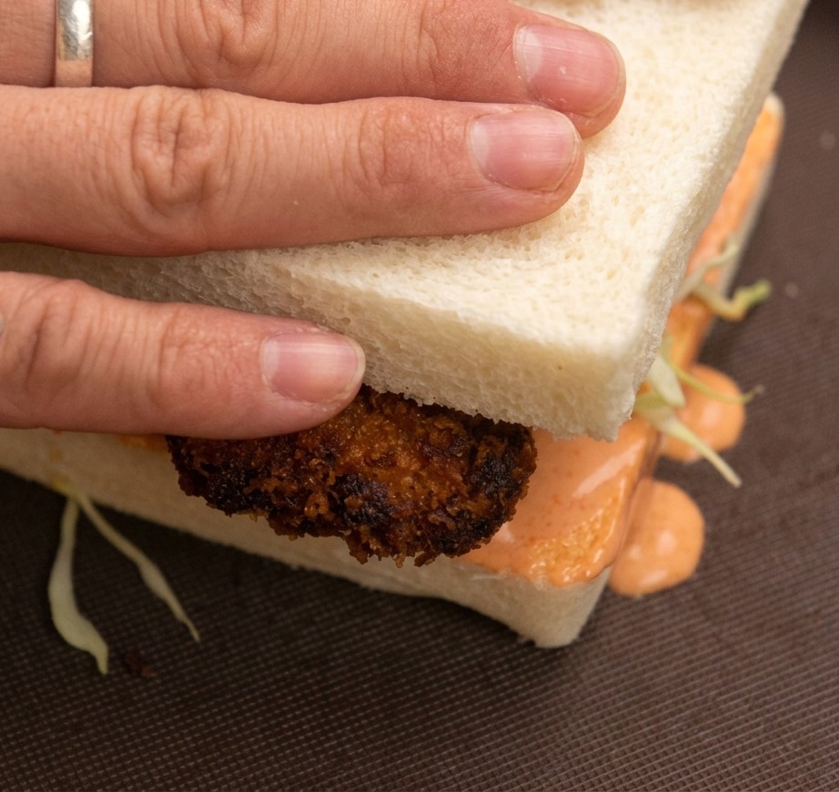 Manos presionando la rebanada superior de un sándwich de pollo frito, con col y mayonesa visibles.