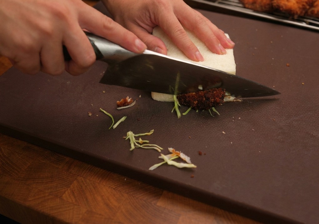 Un cuchillo de cocina grande cortando un sándwich de pollo frito grueso sobre una tabla de madera.