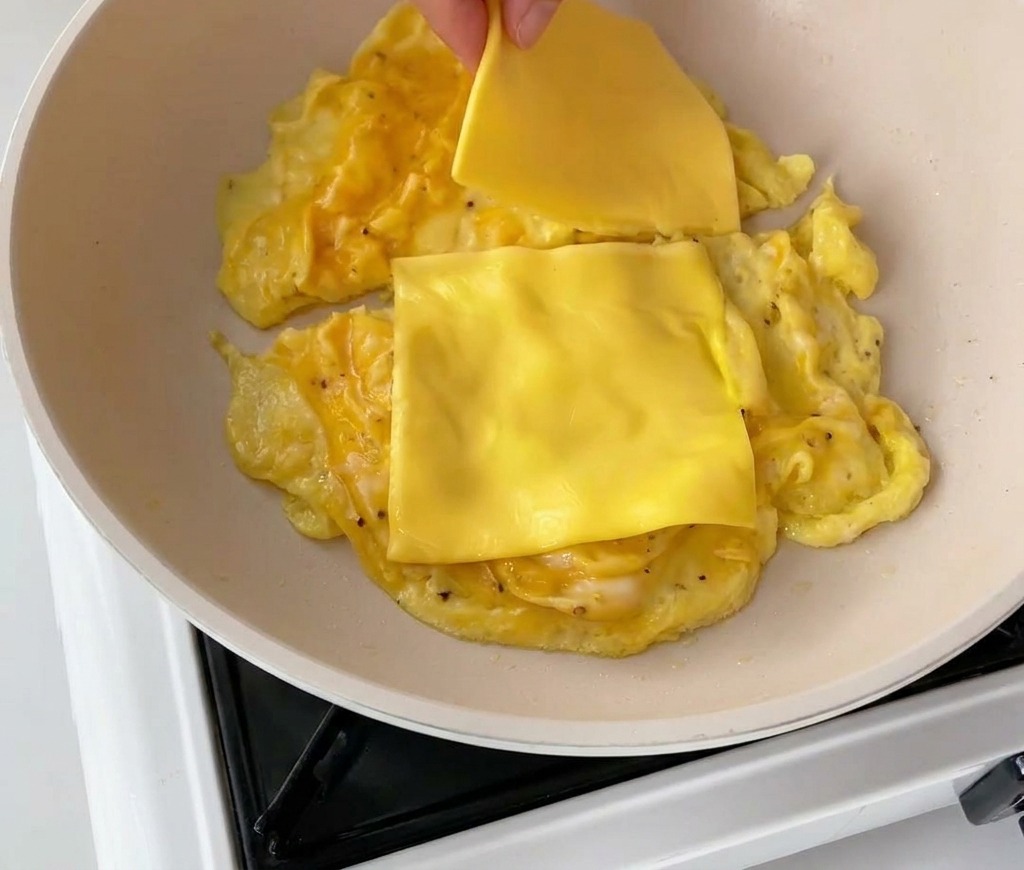 Una mano colocando dos lonchas de queso cheddar amarillo sobre porciones de huevos revueltos.