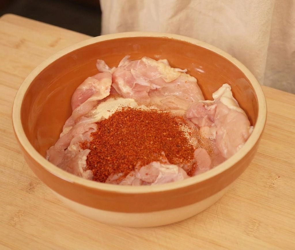 Trozos de pollo crudo en un bol con chile en polvo y condimentos.