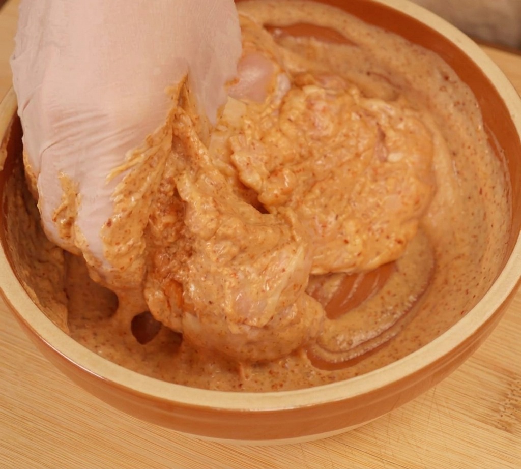 Una mano con guante mezclando trozos de pollo crudo en un marinado de yogur de color naranja.