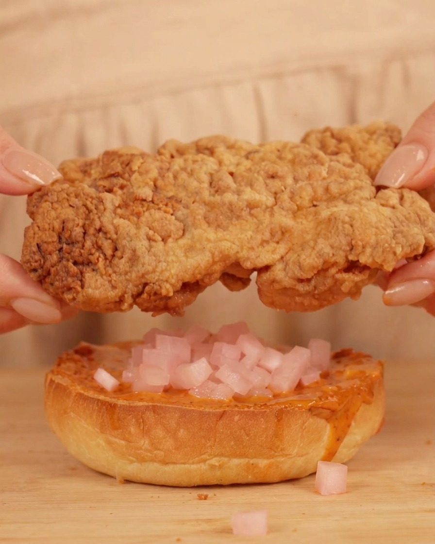 Manos colocando un filete de pollo frito sobre un pan con salsa y rábano encurtido.