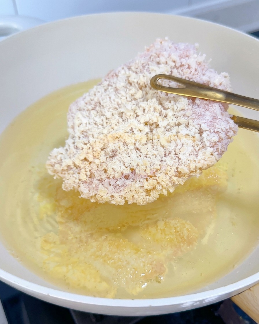 Una milanesa de pollo congelada siendo introducida en aceite caliente con pinzas doradas.