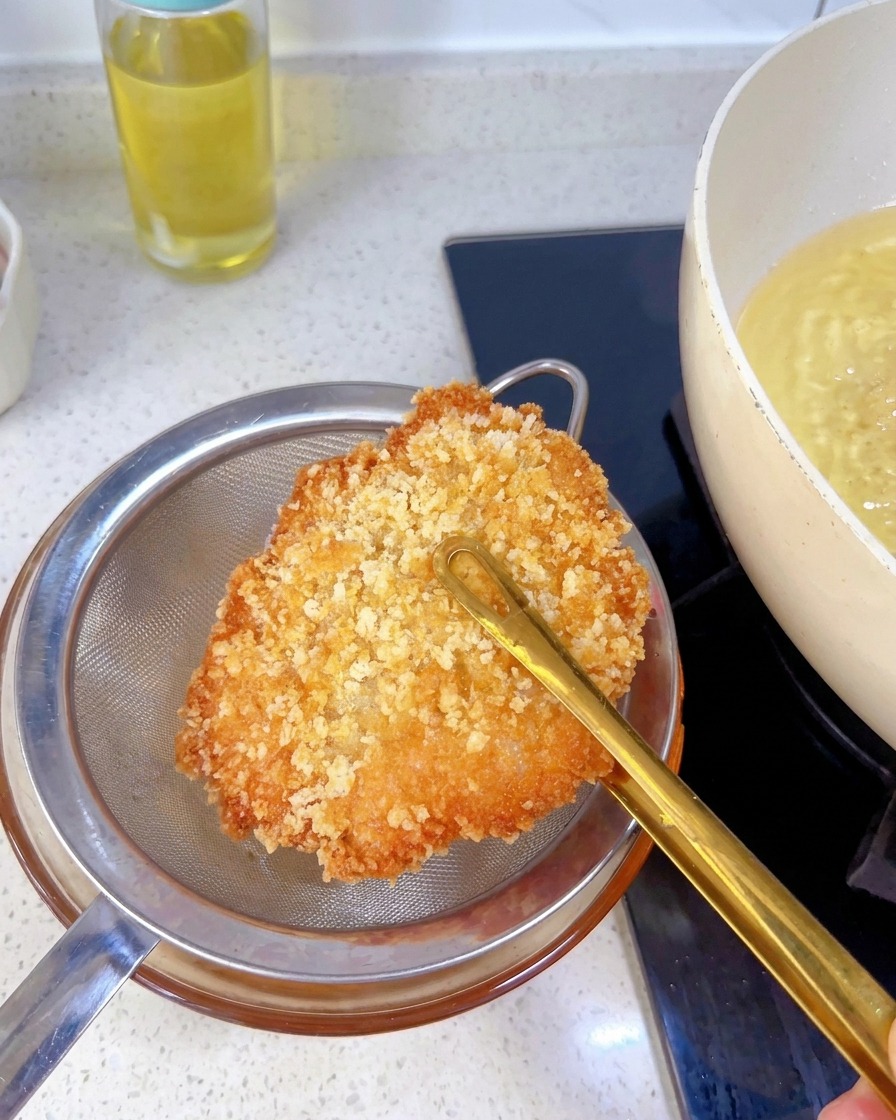 Pollo frito dorado sostenido con pinzas sobre un colador metálico para eliminar el exceso de aceite.