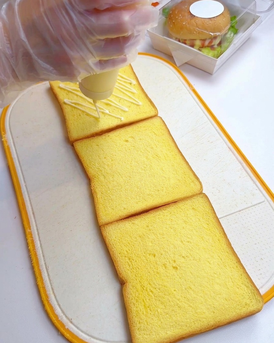 Una mano con guante aplicando mayonesa en zigzag sobre tres rebanadas de pan de molde.