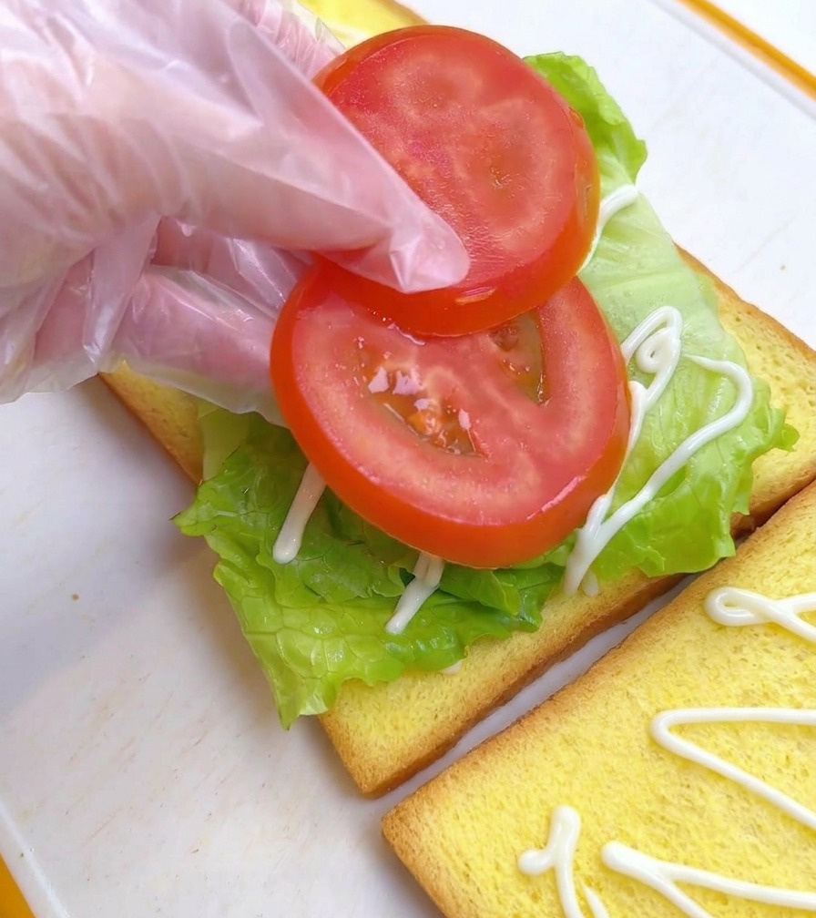 Dedos colocando dos rodajas de tomate rojo sobre la lechuga en el pan de molde.