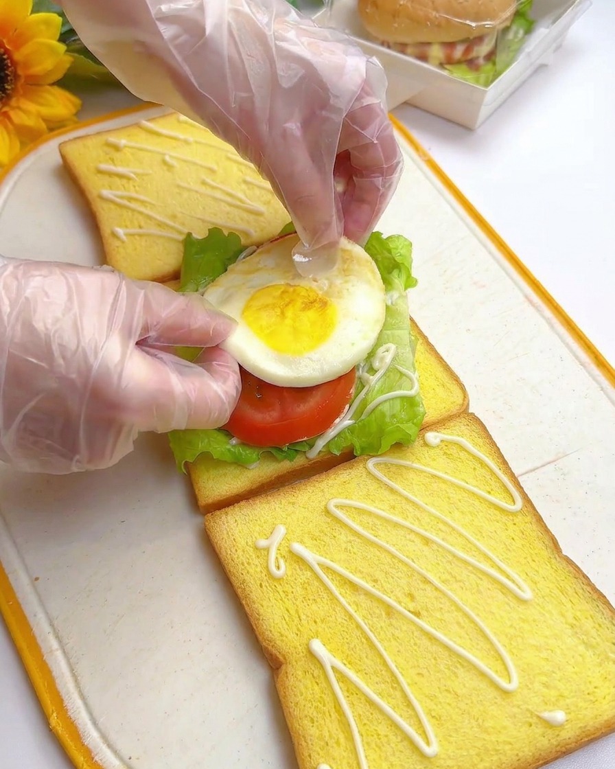 Manos colocando un huevo frito sobre rodajas de tomate y lechuga en el sándwich.