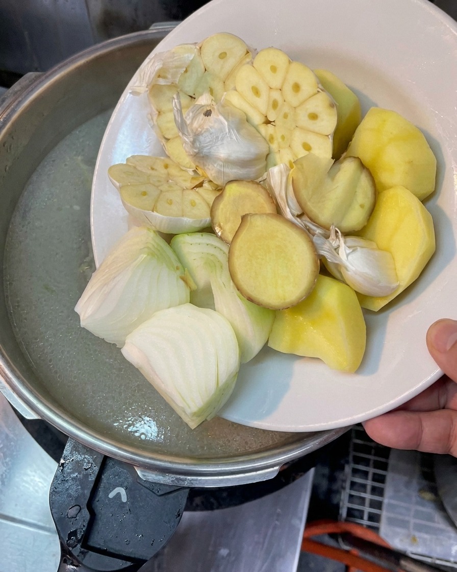 Un plato con cabezas de ajo cortadas a la mitad, cebollas, jengibre y trozos de papa sostenido sobre una olla de caldo.