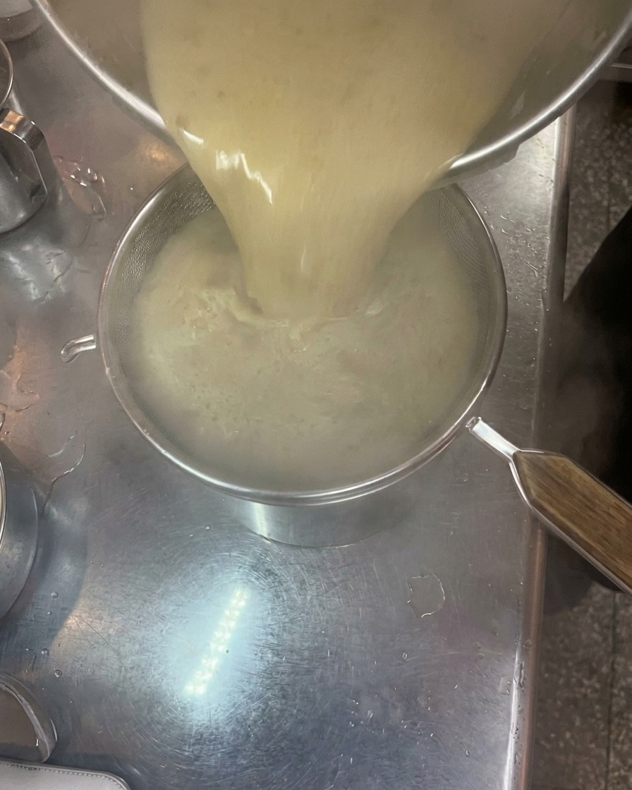 Caldo tonkotsu espeso y cremoso vertiéndose desde una olla a través de un colador fino hacia otro recipiente.