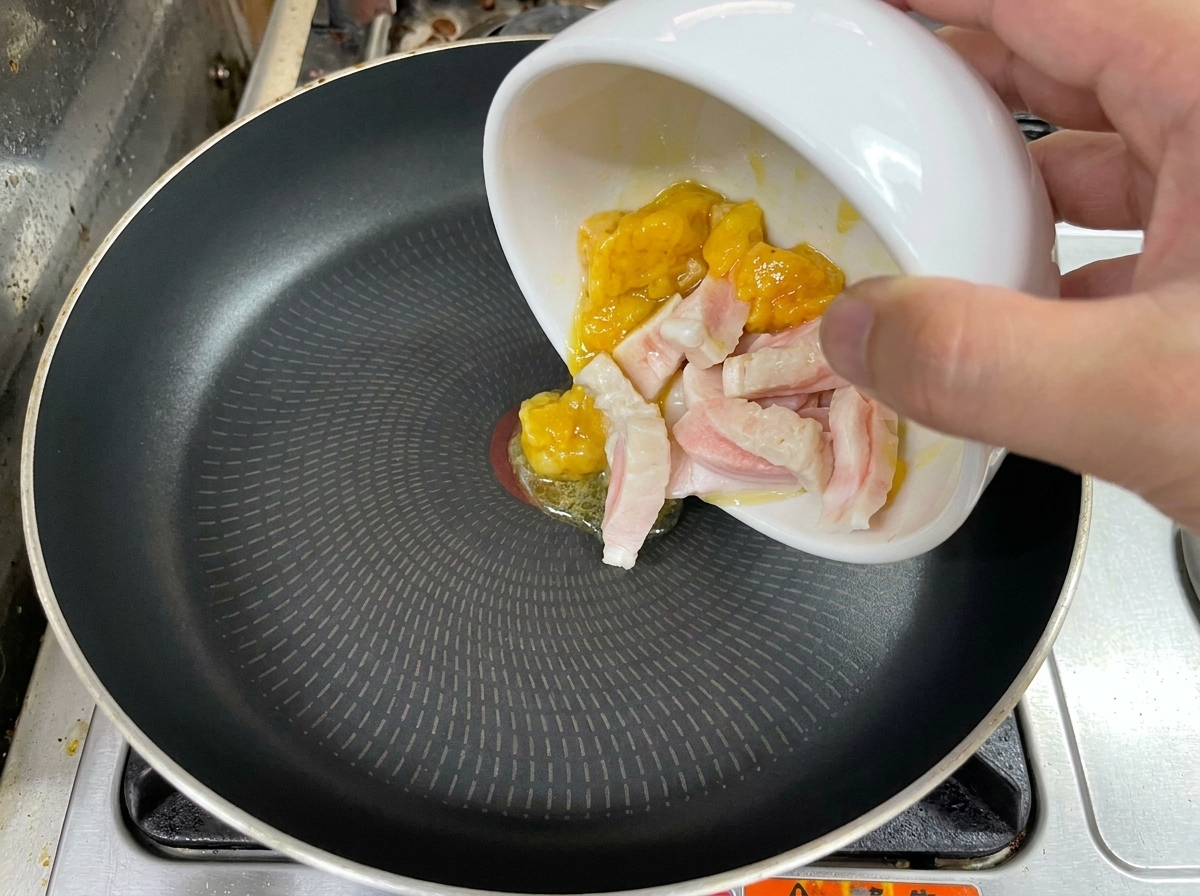 Una mano vertiendo trozos de grasa de pollo y cerdo cruda desde un tazón blanco a una sartén negra.