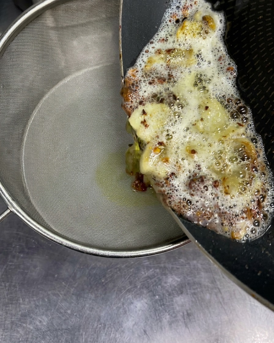 Aceite aromático caliente burbujeando con trozos de ajo frito vertiéndose desde una sartén a un colador fino.