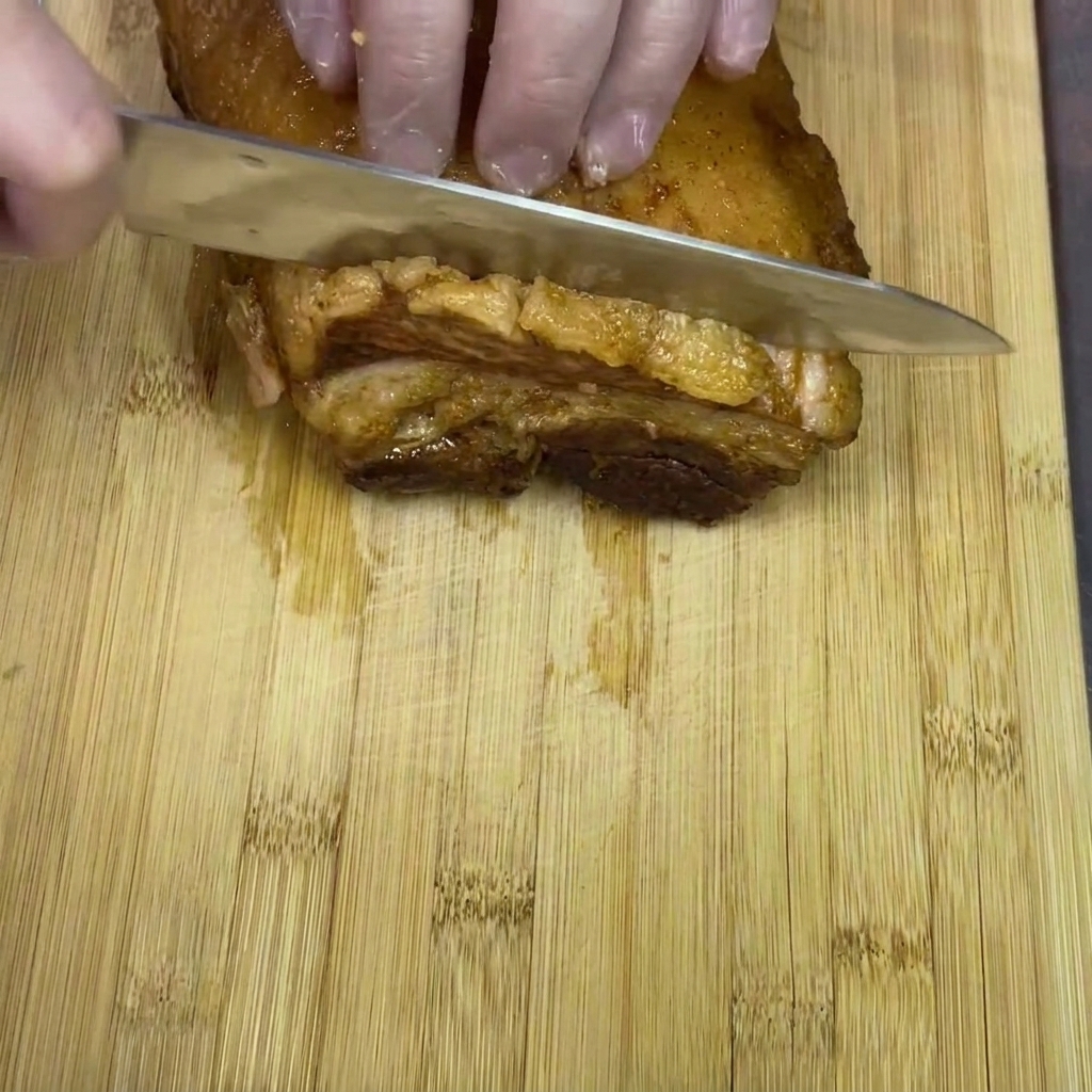 Manos con guantes cortando un bloque de panceta de cerdo braseada en trozos gruesos sobre una tabla de madera.