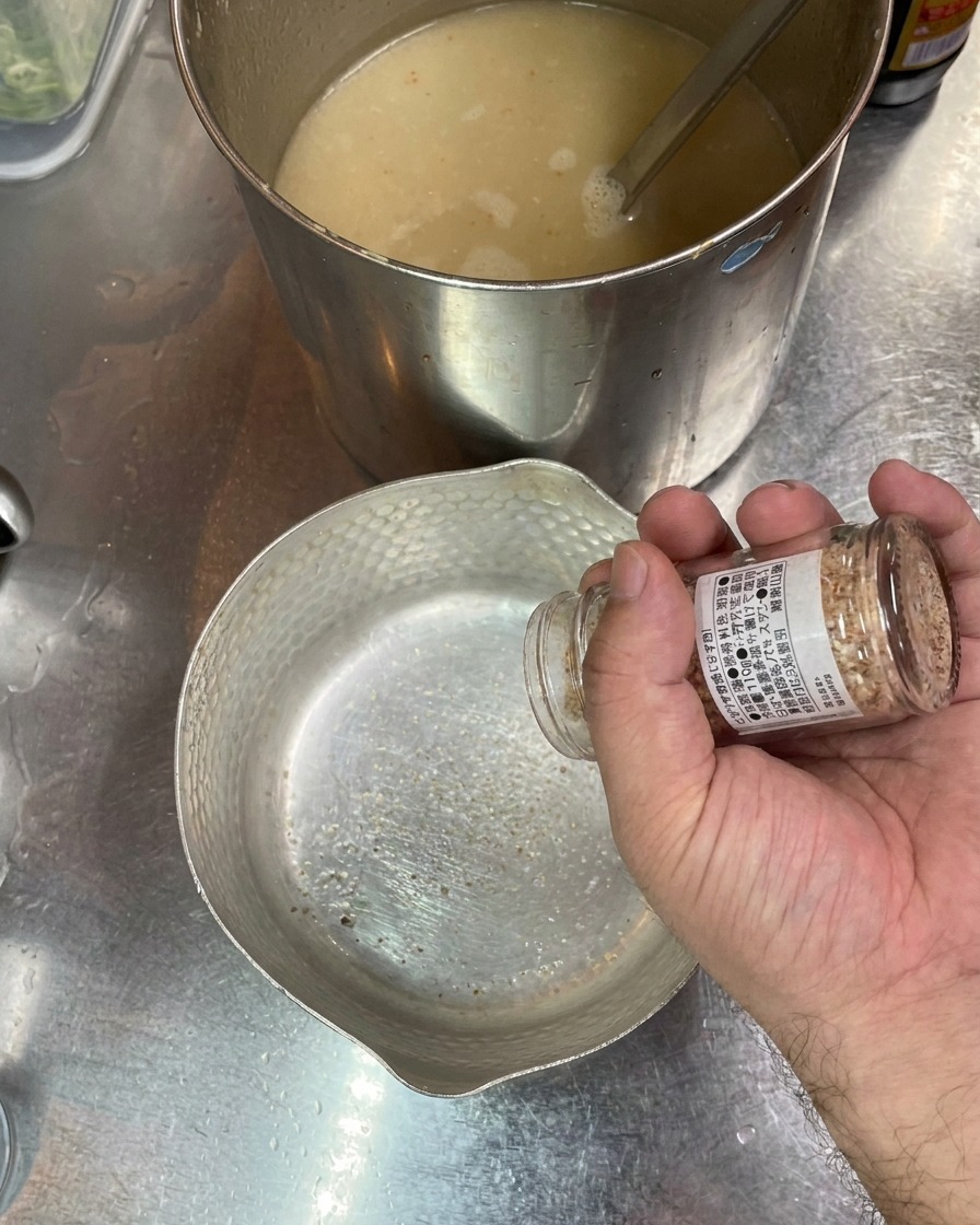 Una mano agitando sal desde un frasco hacia un tazón de metal vacío con una olla de caldo al fondo.