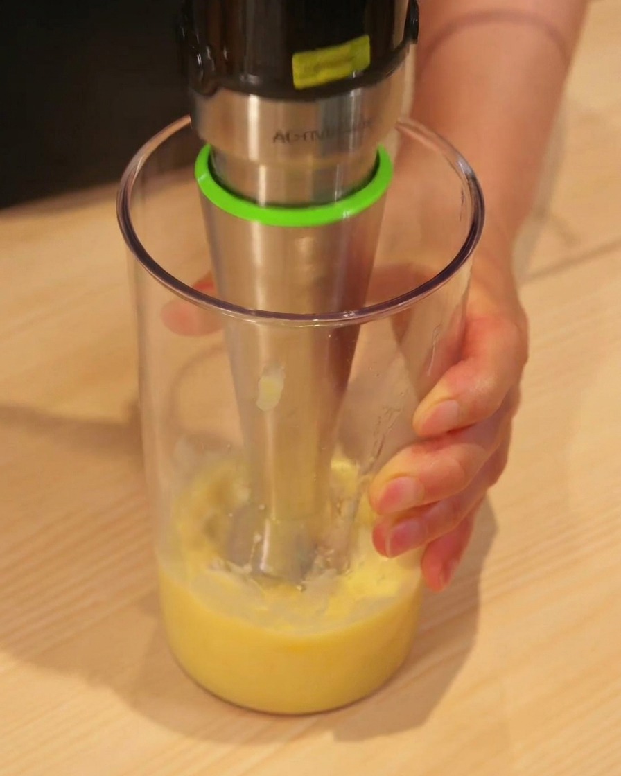 Batidora de mano triturando trozos de piña hasta obtener zumo en un vaso alto.