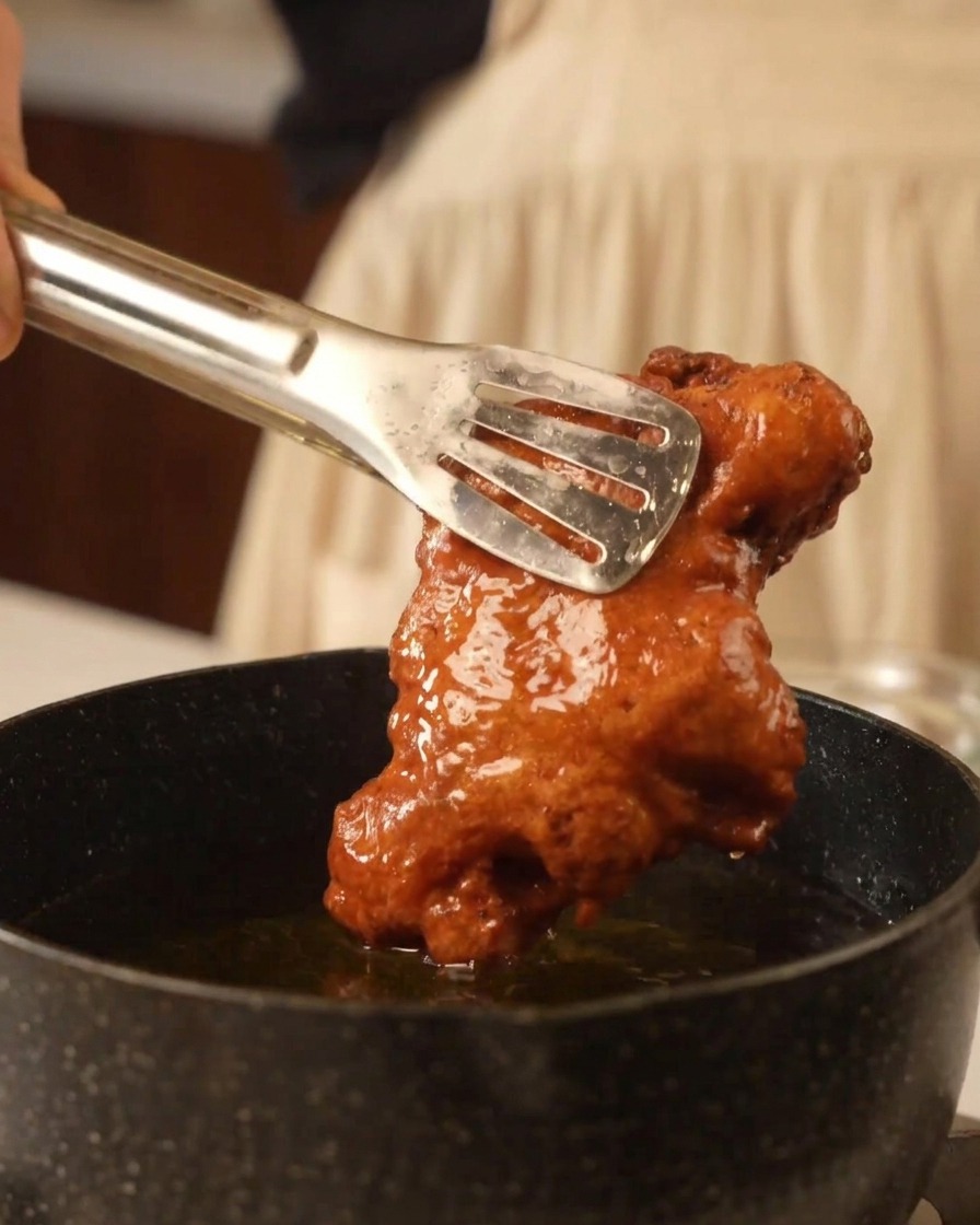 Pinzas de metal levantando un trozo de pollo frito extra crujiente y dorado de una sartén con aceite.