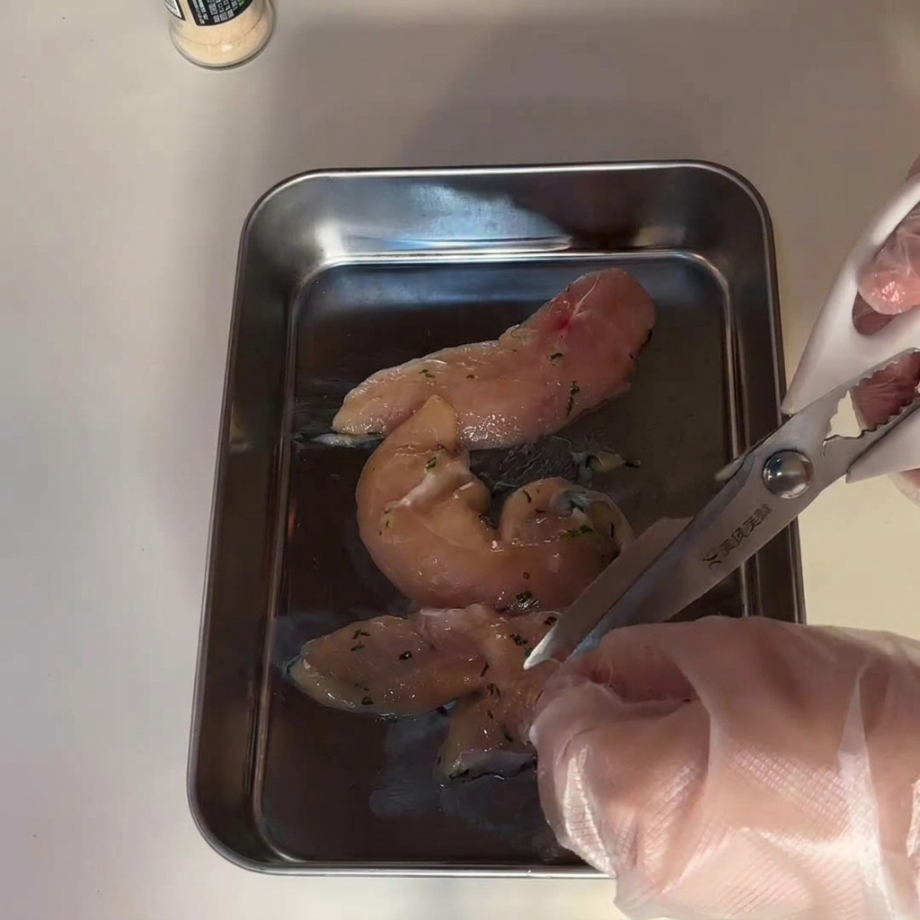 Manos con guantes usando tijeras de cocina para cortar filetes de pechuga de pollo cruda en una bandeja de acero.