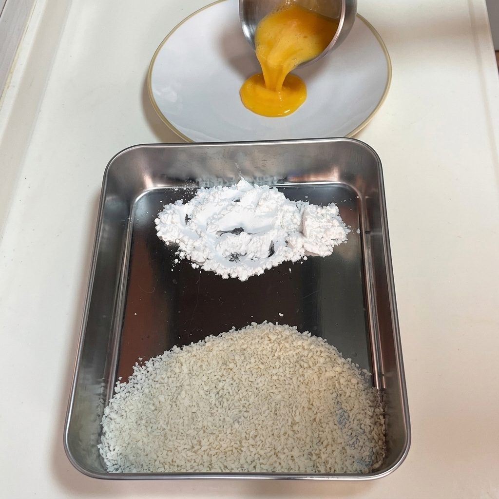 Vertiendo huevo batido en un plato blanco junto a una bandeja con montones separados de maicena y panko.