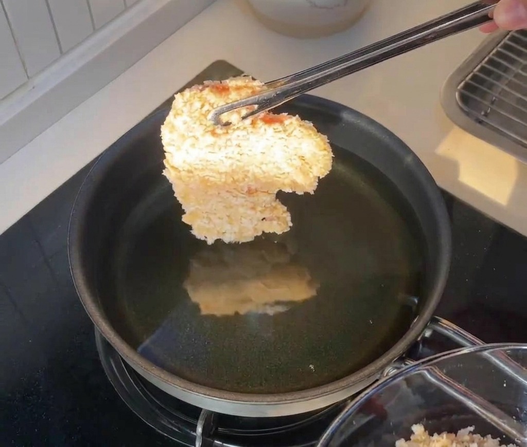 Un filete de pollo rebozado sostenido con pinzas justo encima de una sartén con aceite caliente.
